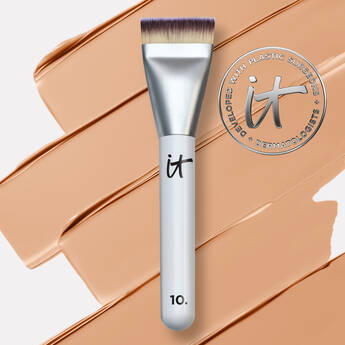 IT Cosmetics makeup brush with flat top bristles placed over tan foundation swatch and brand logo in background.