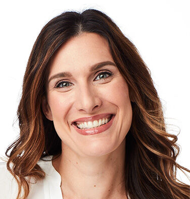 Smiling woman with long brown hair wearing a white top, looking directly at the camera.