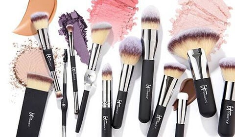 Collection of IT Cosmetics makeup brushes arranged over pressed powder and cream makeup swatches.