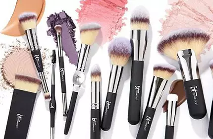 Collection of IT Cosmetics makeup brushes arranged over pressed powder and cream makeup swatches.